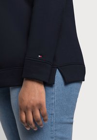 Granatowy sweter z ribbowymi mankietami, bocznymi rozcięciami i małym logo. W zestawieniu z jasnoniebieskimi dżinsami, z ręką opartą obok.