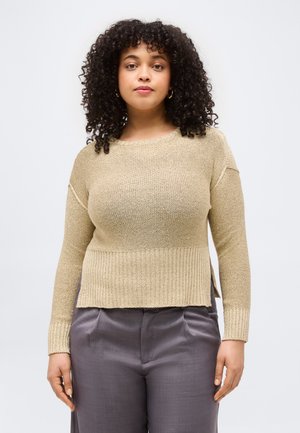 Frau mit lockigem Haar, die einen goldfarbenen Strickpullover und graue Hosen trägt, steht vor einem schlichten hellen Hintergrund.