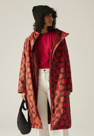 Kobieta w czerwonym i pomarańczowym płaszczu w groszki, na czerwonej koszuli i białych spodniach, trzymająca czarną torebkę, patrzy w bok.