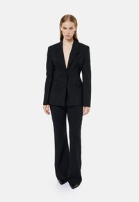 Sort skræddersyet blazer med dyb V-hals, enkelt knaplukning og to forlommer; parret med højtaljede, flagrende sorte bukser.
