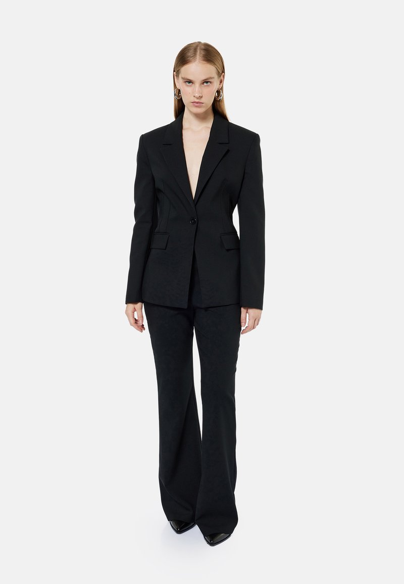 Sort skræddersyet blazer med dyb V-hals, enkelt knaplukning og to forlommer; parret med højtaljede, flagrende sorte bukser.
