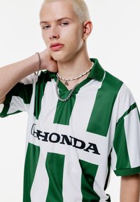 Groen en wit gestreept polo-shirt met korte mouwen en een kraag, met een opvallend zwart "Honda" logo over de borst. Zilveren kettingaccessoires zichtbaar.