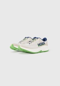 Chaussures de course Hoka avec une tige en mesh respirant beige clair et des accents bleu marine. Semelle extérieure épaisse verte pour l'amorti, avec un logo bien visible.