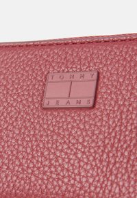 Raudona tekstūruota odinė piniginė su kvadratiniu įspaustu logotipu, kuriame užrašas "TOMMY JEANS" blokiniais rašmenimis. Artimas medžiagos detalių vaizdas.
