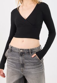 Woman wearing a black ribbed long-sleeve cropped top and high-waisted faded gray jeans with one hand in pocket.