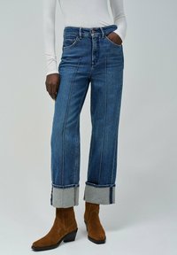 Trenutno visoke plave traperice sa širokim nogavicama, kontrastnim šavovima i prevrnutim manžetama, uparene s smeškim nubuk čizmama do gležnja.