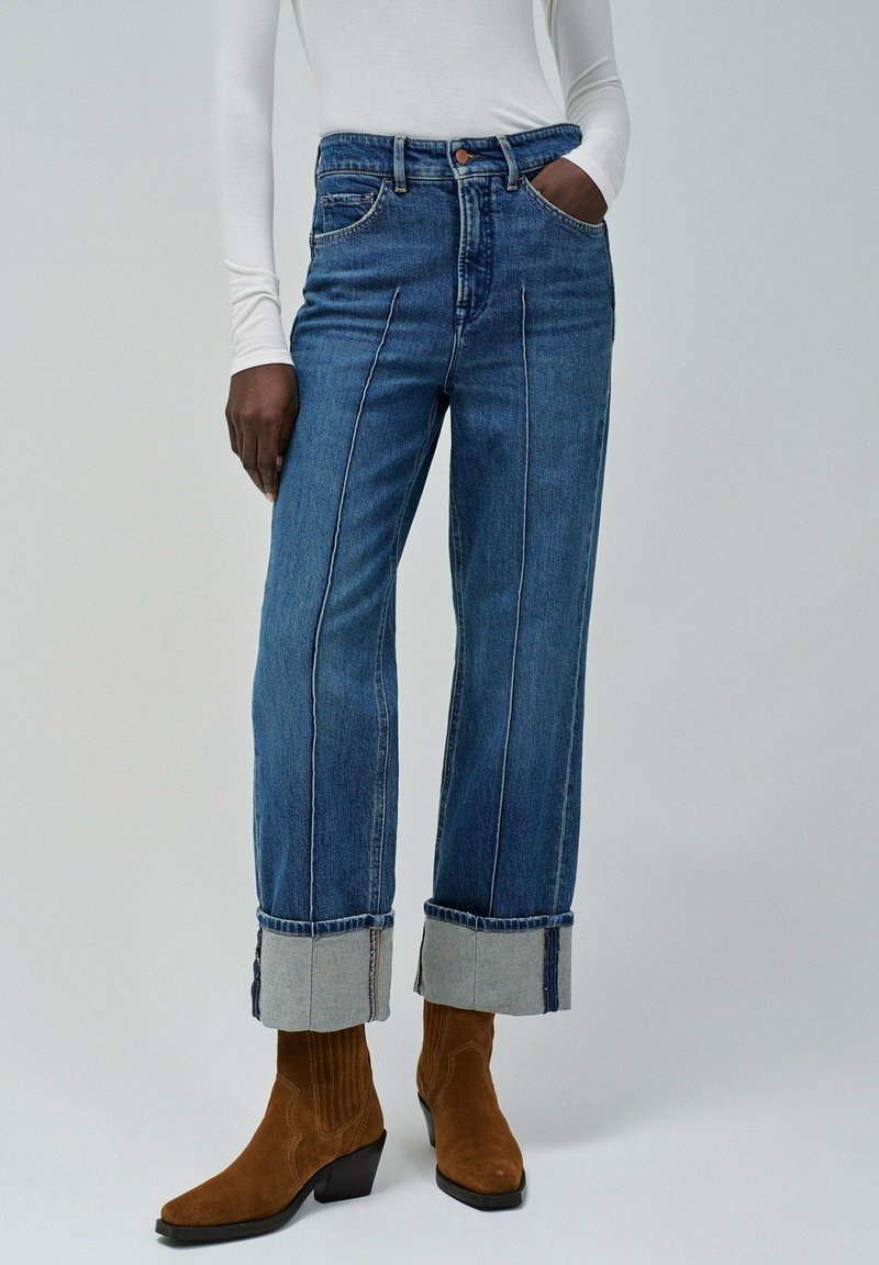 Trenutno visoke plave traperice sa širokim nogavicama, kontrastnim šavovima i prevrnutim manžetama, uparene s smeškim nubuk čizmama do gležnja.