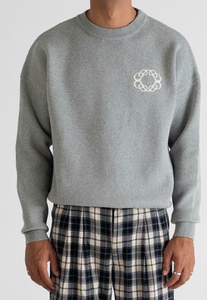Mann trägt einen hellgrauen Strickpullover mit kleinem weißem Ring-Design auf der Brust und schwarz-weiß karierten Hosen.