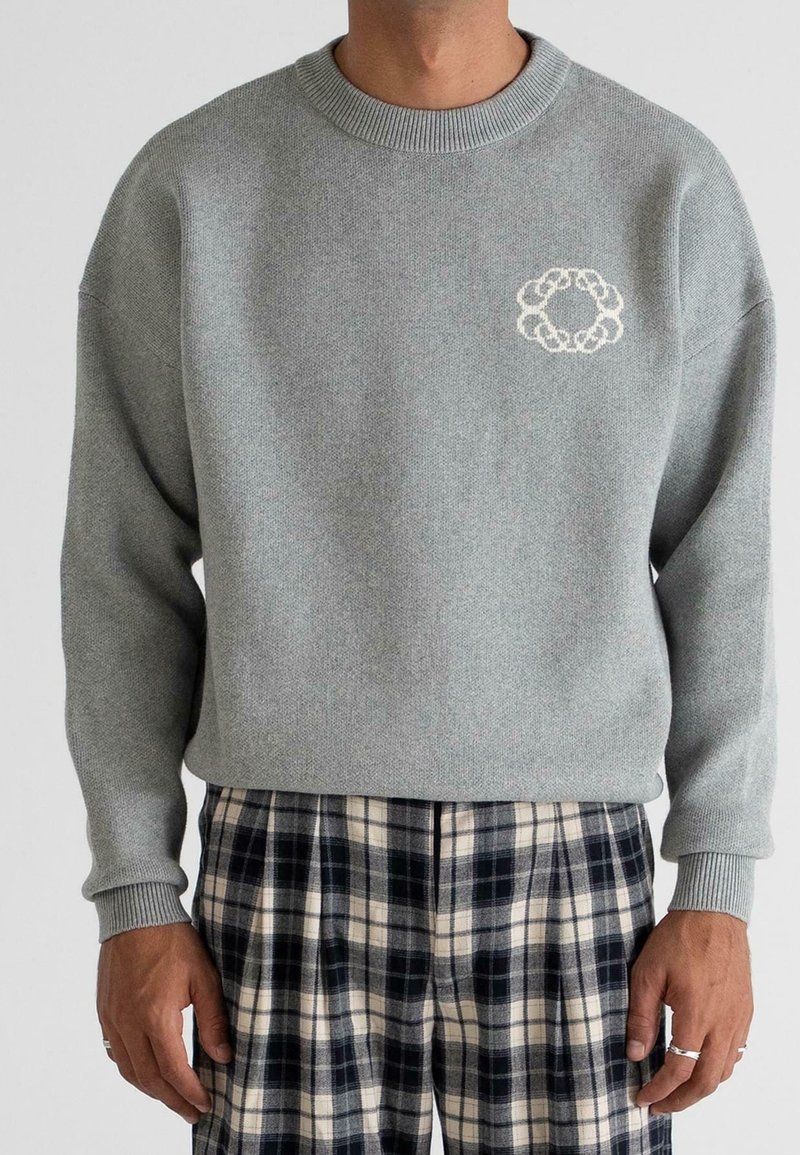 Homme portant un pull en maille gris clair avec un petit motif de cercles blancs entrelacés sur la poitrine et un pantalon à carreaux noir et blanc.