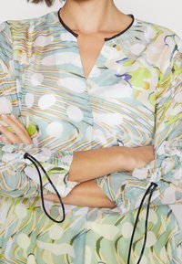 Leichte, gemusterte Bluse in Aquamarin- und Pastellnuancen. Sie verfügt über einen V-Ausschnitt, geraffte Ärmel mit Zugbändern und ein fließendes Design.