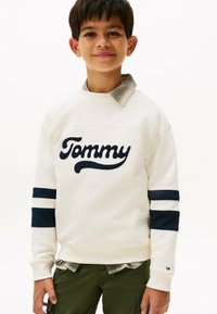 Witte sweatshirt met marineblauwe strepen, met 'Tommy' geborduurd in cursief. Gelaagd over een grijs en wit geruit shirt. Groene cargo broek.