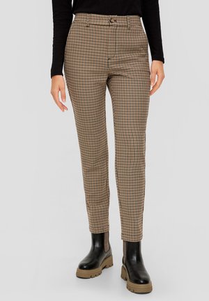 Femme portant un pantalon marron et noir à motif pied-de-poule avec bouton sur le devant et des bottes noires à semelle épaisse, debout devant un fond uni.