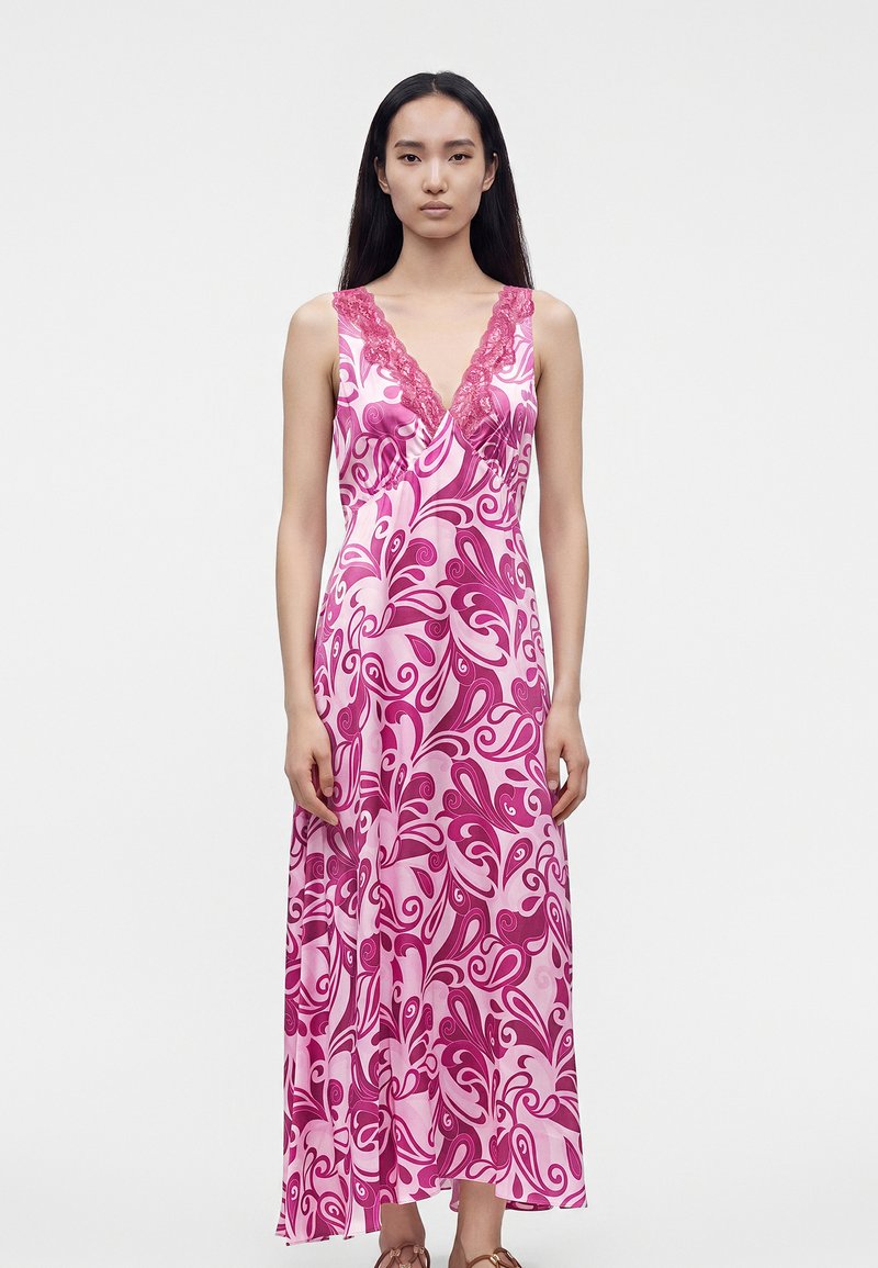 Femme portant une robe sans manches à motif floral rose et violet avec bordure en dentelle au col en V, debout devant un fond blanc uni.