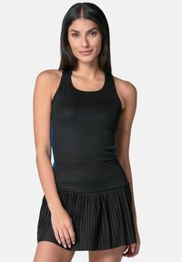 Vrouw met lang, donker haar, gekleed in een mouwloze zwarte top en een geplisseerde zwarte rok, staande tegen een effen lichte achtergrond.