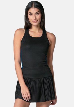 Vrouw met lang, donker haar, gekleed in een mouwloze zwarte top en een geplisseerde zwarte rok, staande tegen een effen lichte achtergrond.