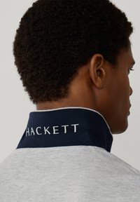 Muž s krátkymi kučeravými vlasmi oblečený v sivej košeli s tmavomodrým stojacím golierom, na ktorom je bielym písmom nápis "Hackett".