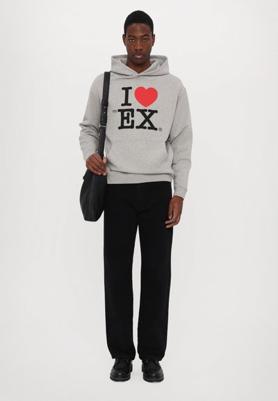 Felpa grigia con cappuccio e stampa grafica rossa e nera con la scritta "Amo il mio EX", abbinata a pantaloni neri e scarpe nere, completata da una borsa nera.