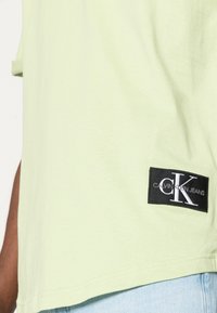 T-shirt vert clair avec un patch logo Calvin Klein Jeans noir près de l'ourlet, porté avec un jean bleu clair.