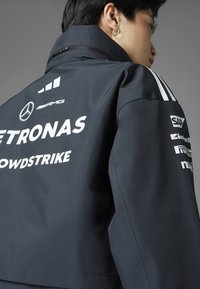 Crna vodootporna jakna s bijelim logotipima, uključujući Mercedes i Petronas. Dizajnirana s visokim ovratnikom i kontrastnim prugama na ramenima.