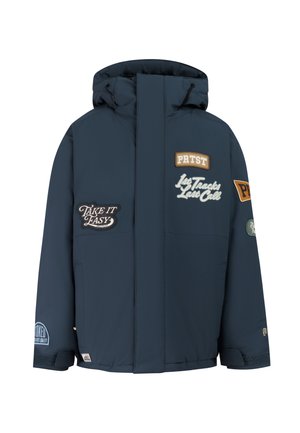 Jonge persoon in donkerblauwe winterjas met patches, beige broek, zwarte sneeuwlaarzen, beige muts en zwarte skibril op het hoofd.