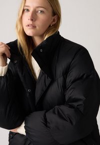 Juoda pūkinė striukė su aukštu apykakliu, spaudėmis ir minkšta pamušalu viduje. Turi dygsniuotą tekstūrą ir laisvą, per didelį kirpimą.