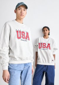 Felpe grigio chiaro a girocollo con grafica rossa "USA" e testo "Tommy Jeans". Entrambi gli individui indossano jeans di denim blu con una vestibilità rilassata.