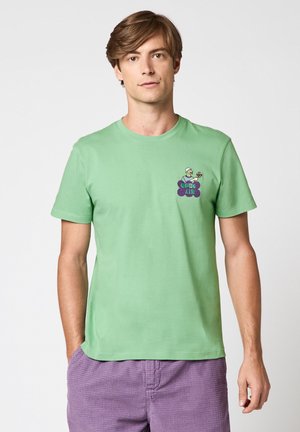 Jeune homme portant un t-shirt vert clair avec un petit motif et le texte « OPEN AIR » sur la poitrine, assorti à un short violet texturé.