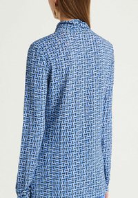 Chemise à manches longues en tissu bleu avec un motif géométrique dans des tons plus clairs. Elle possède un col montant et une texture douce avec une coupe ajustée.