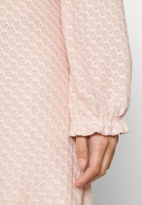 Vestido rosa claro com padrão texturizado em favo de mel, apresentando mangas longas e punhos franzidos. Um detalhe de tecido suave é visível perto da mão.