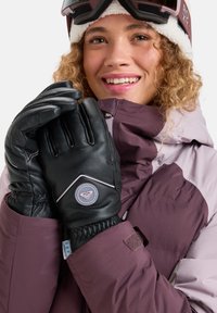 Lächelnde Person, die Skibrillen, eine lila Winterjacke und schwarze Handschuhe trägt und die Hände nah ans Gesicht hält, vor einem weißen Hintergrund.
