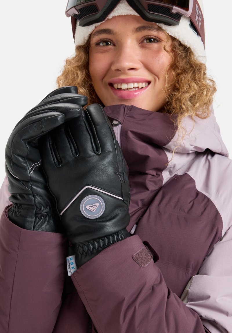 Lächelnde Person, die Skibrillen, eine lila Winterjacke und schwarze Handschuhe trägt und die Hände nah ans Gesicht hält, vor einem weißen Hintergrund.