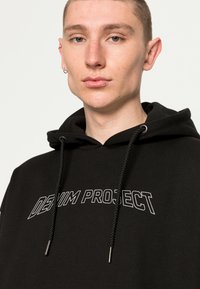 Felpa nera in cotone con il logo "DENIM PROJECT" in bianco. Include un cordino con punte metalliche e una tasca a marsupio.
