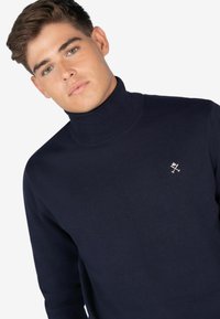 Maglione a collo alto navy realizzato in un tessuto a maglia liscio con una sottile lucentezza. Presenta un piccolo logo ricamato sulla parte superiore sinistra del petto.