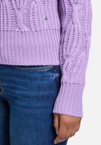 Helllila gestrickter Pullover mit einem tiefen Rippenbund und Zopfmuster; getragen mit dunkelblauen Jeans, die detaillierte Nähte zeigen.