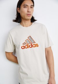 Tricou din bumbac bej, cu un logo mare portocaliu "adidas", accente texturate în cadrul logo-ului și un gât rotund. Mâneci scurte.