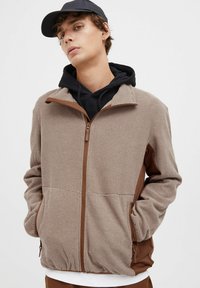 Brun zip-up jakke med teksturert stoff og kontrasterende brune detaljer. Har sidelommer og er lagdelt over en svart hettegenser.