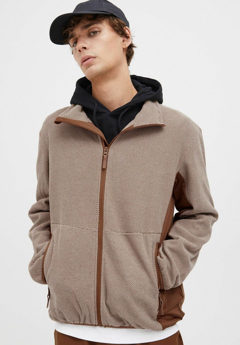 Brun zip-up jakke med teksturert stoff og kontrasterende brune detaljer. Har sidelommer og er lagdelt over en svart hettegenser.