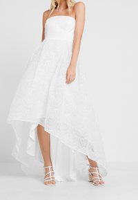 Robe blanche sans bretelles à ourlet asymétrique, avec une superposition en dentelle texturée et une doublure intérieure lisse. Portée avec des talons blancs à lanières.