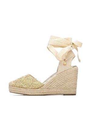 Alpargata de cuña con parte superior de tela beige texturizada con bordados intrincados, acabado con un tacón de yute tejido y una correa para atar alrededor del tobillo.