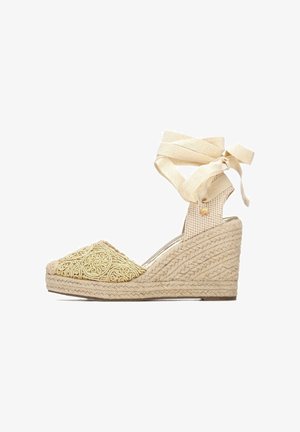 Alpargata de cuña con parte superior de tela beige texturizada con bordados intrincados, acabado con un tacón de yute tejido y una correa para atar alrededor del tobillo.