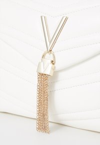 Borsa a tracolla in pelle bianca trapuntata con una chiusura a forma di V dorata e una nappina a catena. La chiusura presenta un piccolo dettaglio inciso a forma di V.