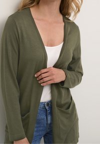 Olivově zelený kardigan s volnějším střihem, otevřený vpředu a se dvěma kapsami, kombinovaný s bílým tílkem a modrými džíny, s jemnou texturou.