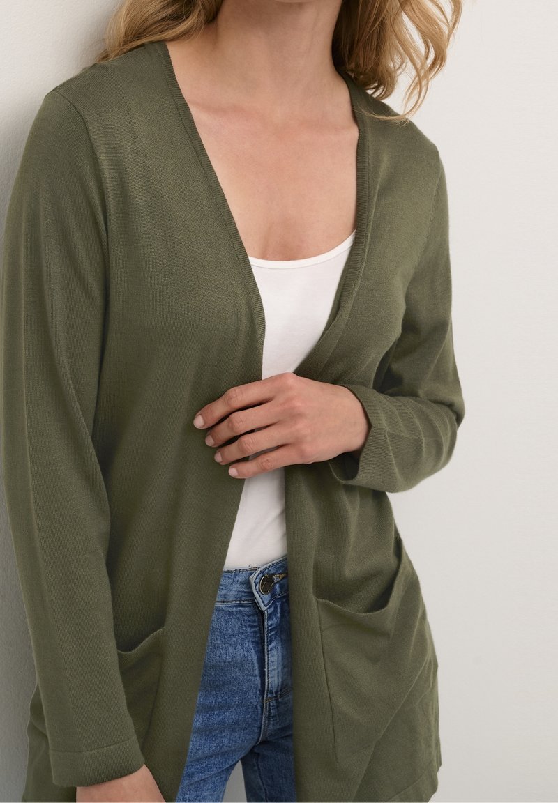 Olivově zelený kardigan s volnějším střihem, otevřený vpředu a se dvěma kapsami, kombinovaný s bílým tílkem a modrými džíny, s jemnou texturou.