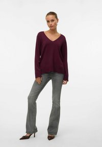 Maglione bordeaux con scollatura a V e maniche lunghe, abbinato a jeans grigi chiari a zampa d'elefante e tacchi a punta marroni, su uno sfondo bianco.