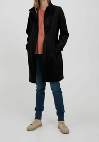 Cappotto nero fino al ginocchio con colletto alto, chiusura con zip e tasche laterali, abbinato a una maglietta corallo e jeans blu, completato da sneakers beige.