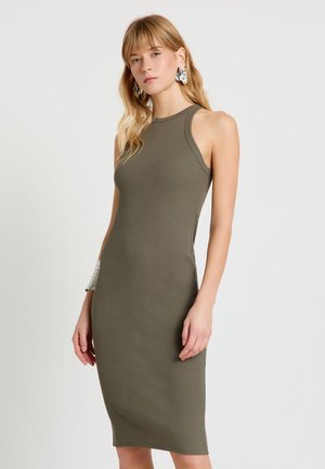 Giovane donna con lunghi capelli biondi, indossa un abito aderente senza maniche di colore verde oliva e grandi orecchini d'argento, su uno sfondo chiaro e uniforme.