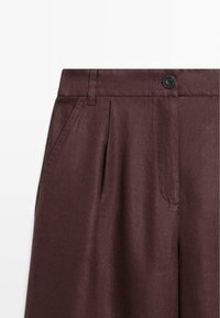 Gros plan d’un pantalon marron foncé avec poches latérales, plis, passants de ceinture et un bouton noir à la taille.