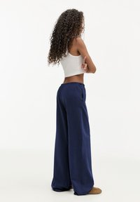 Kvinde med langt krøllet hår iført en hvid ærmeløs crop top, marineblå brede bukser og brune sko, stående med armene korslagt og vendt sidelæns.