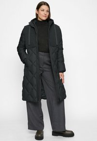 Woman wearing long black quilted coat with textured upper panel, black turtleneck, gray wide-leg pants, and platform boots, standing against white background.