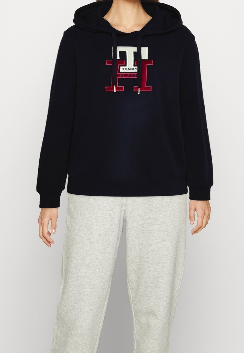 Personne portant un sweat à capuche noir avec un grand logo "TH" rouge et blanc et le texte "Tommy Hilfiger", associé à un pantalon de survêtement gris clair.